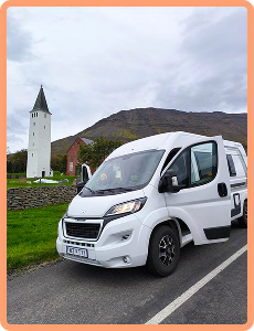 Camping-car compact blanc garé juste à côté d'un clocher dans une zone reculée d'Islande.